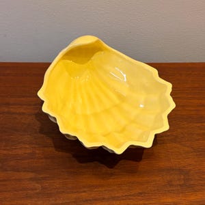 May include: A yellow ceramic seashell-shaped dish. The dish has a glossy finish and a scalloped edge. The interior of the dish has a textured, fan-like pattern. The dish is sitting on a dark wooden surface.