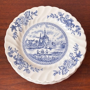May include: A stack of white ceramic plates featuring a blue and white design. Each plate has a central image of a windmill and figures near water, surrounded by floral patterns and a scalloped edge.