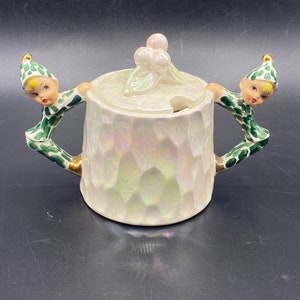 May include: White ceramic sugar bowl with a lid featuring two green and gold elves holding the handles. The lid has a small pink flower design.