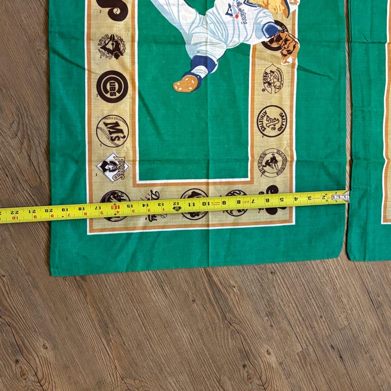 May include: Green fabric wall hanging with a baseball player in a white uniform and a brown baseball cap. The fabric has a tan border with logos of different baseball teams, including the Cubs, Twins, and Pirates.