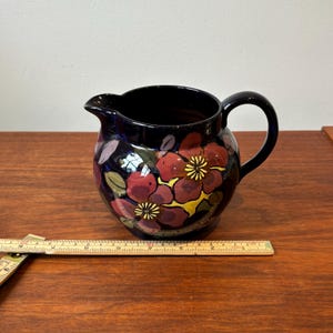 Op de afbeelding: Een donkerblauwe keramische kan met een handvat en tuit. De kan is versierd met rode en gele bloemmotieven en groene bladeren. De kan is ongeveer 15 cm hoog.