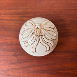 May include: A circular, cream-coloured ceramic trinket box with a gold bow design on the lid. The bow has detailed gold outlines and accents, creating a decorative and elegant appearance. The box is set against a warm, wooden surface.
