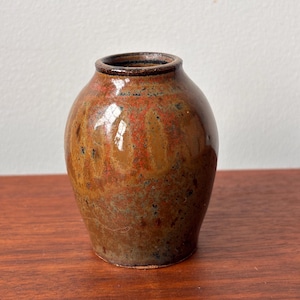 May include: A small, handmade ceramic vase with a rounded form and a narrow neck. The vase has a mottled glaze in shades of brown, red, and green. The rim is a darker brown. The vase is on a wooden surface.
