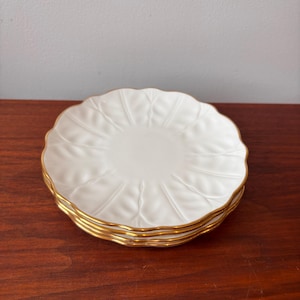 May include: A stack of four white ceramic plates with a gold rim. Each plate has a scalloped edge and a leaf-like embossed design radiating from the center. The plates are stacked on a dark wooden surface.
