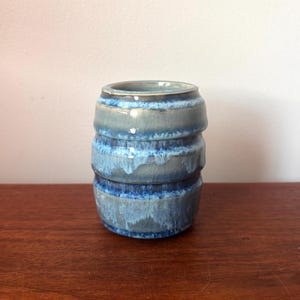 May include: A handcrafted ceramic vase with a stacked barrel design. The vase features a gradient of blue hues, from light to dark, with a textured, crackled glaze. The interior is a darker blue, and the vase sits on a wooden surface.