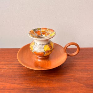 Puede incluir: Un jarrón de cerámica vintage con un diseño floral en naranja, amarillo y verde se encuentra sobre un plato de madera con asa. El jarrón tiene una base color crema y una parte superior acampanada. El plato de madera es redondo y de tono marrón rojizo cálido.