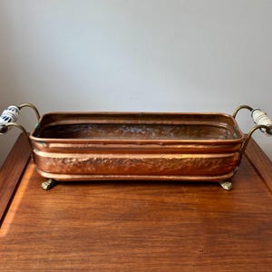 May include: A rectangular hammered copper planter with a metallic finish, featuring two curved brass handles on each side. The handles are accented with white ceramic grips decorated with blue floral patterns, and the base rests on four small feet.