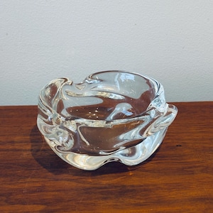 May include: A clear glass ashtray with a flowing, sculptural form. The ashtray features a shallow bowl with undulating edges. It is positioned on a wooden surface. The glass reflects light, producing highlights and shadows.