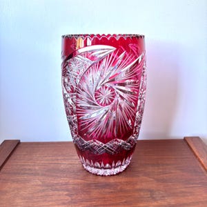 May include: A deep red cut-to-clear crystal vase featuring intricate geometric patterns, including starburst motifs and fan-shaped cuts. The glass displays a vibrant ruby hue with clear, polished facets that create a complex, reflective surface texture.