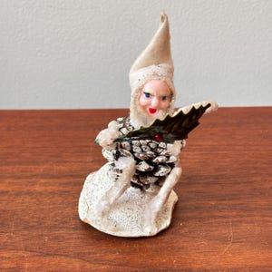 May include: A vintage Christmas figurine of a seated elf with a cone-shaped body and a felt hat. The elf has a painted face, holding a green holly leaf. The figurine is decorated with white glitter and is likely from the mid-20th century.