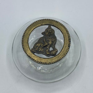 Puede incluir: Un tarro de vidrio transparente con una tapa de latón que presenta una figura de gato detallada. La tapa tiene un diseño decorativo de cuerda alrededor del borde.
