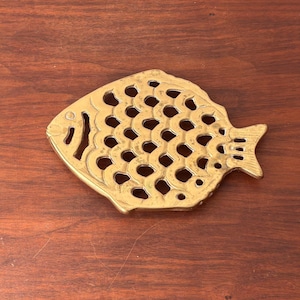 May include: A brass fish-shaped trivet with a decorative openwork design. The trivet is gold-colored and has a detailed fin and tail. The trivet is designed to protect surfaces from hot dishes.