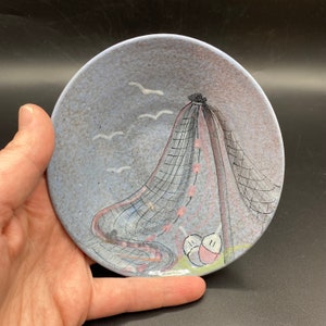 Peut inclure: Une petite assiette ronde en céramique bleue et grise avec un motif peint d'un filet de pêche, deux oiseaux blancs et deux oiseaux roses et blancs. L'assiette a un glaçage moucheté.