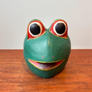 Pode incluir: Uma caixa verde em forma de sapo com um rosto sorridente. O sapo tem olhos grandes e pretos com contornos vermelhos e uma boca rosa. A caixa está sobre uma superfície de madeira.