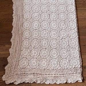 May include: Cream-colored, crocheted blanket with a floral pattern. The blanket features a scalloped edge and a detailed openwork border. The intricate design and neutral color make it a versatile home decor item.