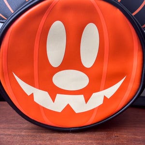 May include: Orange pumpkin-shaped bag with a smiling jack-o'-lantern face. The bag has a black trim and a black handle. The pumpkin face is white with black outlines. A fun Halloween accessory.