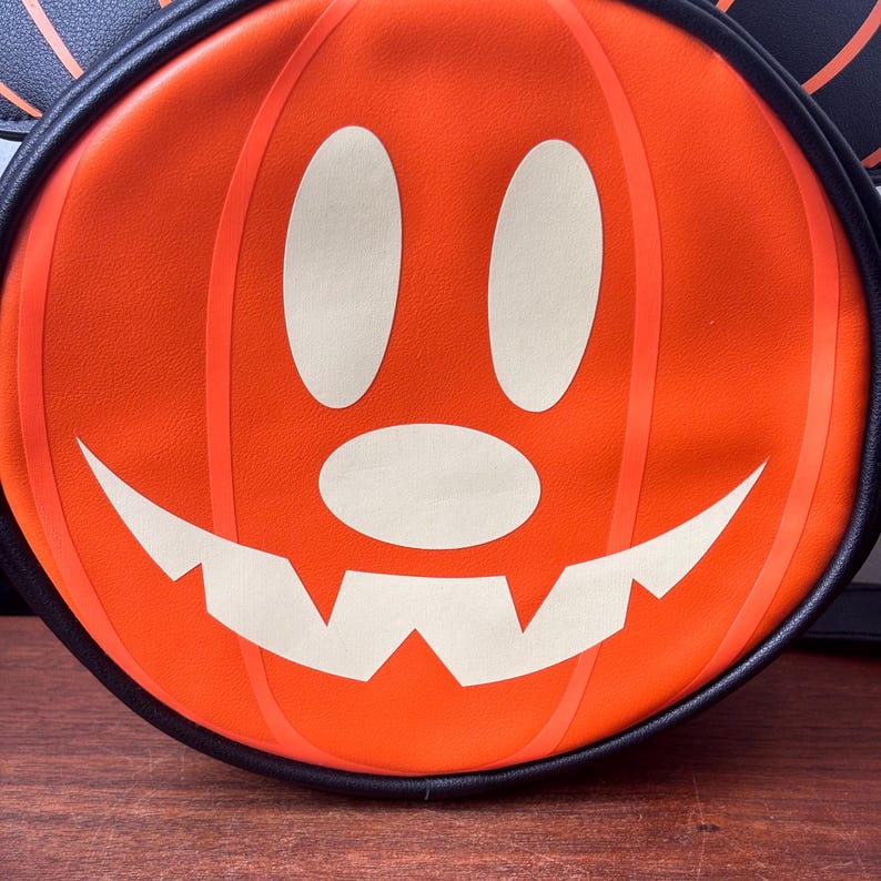 May include: Orange pumpkin-shaped bag with a smiling jack-o'-lantern face. The bag has a black trim and a black handle. The pumpkin face is white with black outlines. A fun Halloween accessory.