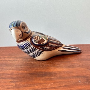 Op de afbeelding: Een keramieken vogelbeeldje met een crèmekleurig lichaam, bruine vleugels en blauwe en groene accenten. De vogel heeft een witte snavel en staat op een houten oppervlak. Het beeldje is een decoratief item.