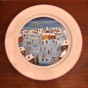 Puede incluir: Plato de cerámica decorativo con una escena invernal. El plato presenta una imagen central de un pueblo nevado con personas, casas y una iglesia. El borde es blanco con una fina raya roja.