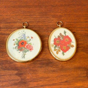 Op de afbeelding: Twee vintage bloemenprints met lijst. Eén is rond en de andere ovaal. Het ronde frame heeft een boeket van rode klaprozen en roze pioenrozen. Het ovale frame toont rode bloemen met groene bladeren.