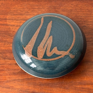 May include: A dark blue ceramic lid with a copper-colored design inside a circle. The design is a stylized abstract shape.