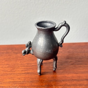 May include: A small, antique pewter creamer with a handle and three legs. The creamer has a bulbous body and a spout with a decorative element. The pewter has a dark, aged appearance, suggesting its vintage.