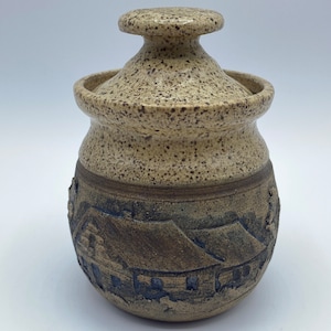 Puede incluir: Un tarro de cerámica con tapa, con un esmalte moteado marrón y beige. El tarro tiene un diseño en relieve de casas con acentos marrones y negros.