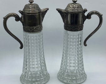 May include: Two vintage glass pitchers with silver-colored metal lids and handles. The clear glass bodies have a textured, diamond-patterned design. The pitchers are tall and slender, with a flared base and a decorative finial on the lid.