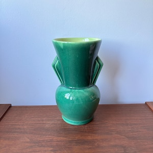 May include: A green ceramic vase with a glossy finish. The vase has a bulbous base, a narrow neck, and a flared top. Two decorative, angular handles are on either side of the neck. The interior is a lighter shade of green.