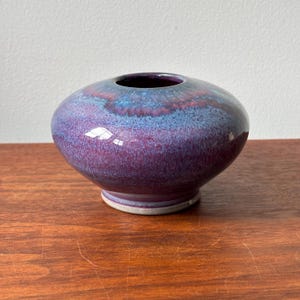 May include: A squat, rounded ceramic vase featuring a glossy glaze with blended shades of deep purple, magenta, and vibrant blue. The vessel has a wide, bulbous body that tapers down to a narrow, light-colored circular base and a small opening.