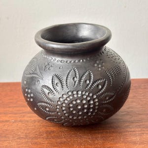 Puede incluir: Una olla de cerámica gris oscuro con intrincados diseños florales y de puntos. La olla hecha a mano tiene un cuerpo redondeado, una abertura ancha y un borde ligeramente ensanchado. La superficie presenta diseños en relieve, creando una apariencia texturizada. La olla está sobre una superficie de madera.
