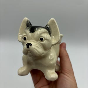 May include: A white ceramic figurine of a French bulldog with black ears and a black nose. The figurine is sitting on its hind legs with its front paws extended.