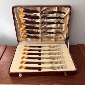 May include: A vintage cutlery set in a brown leather case. The set includes steak knives with brown and yellow handles and forks with matching handles. The knives have serrated blades. The case is lined with a cream-colored satin fabric.
