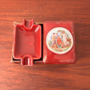 以下が含まれることがあります： ヴィンテージの赤いセラミック灰皿とマッチする蓋。蓋には、金色のフィリグリーで縁取られた庭の人物の絵が描かれています。灰皿は長方形で、曲線的なデザインです。