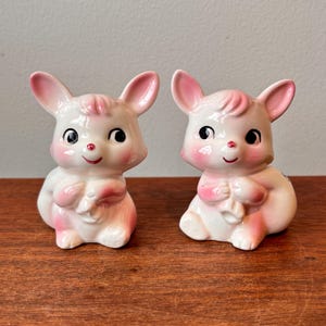 May include: Two ceramic bunny figurines. The bunnies are white with pink accents on their ears, cheeks, and paws. They have black eyes and red noses. The bunnies are sitting and holding a small object. The figurines are on a wooden surface.