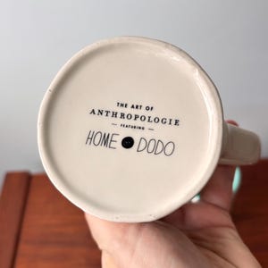 Puede incluir: Base de taza de cer&aacute;mica blanquecina con el texto "THE ART OF ANTHROPOLOGIE - FEATURING - HOME DODO" en negro. La taza tiene un asa y la sostiene una persona.