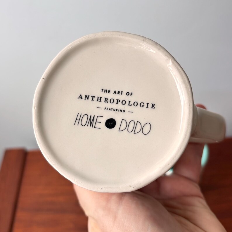 Puede incluir: Base de taza de cer&aacute;mica blanquecina con el texto "THE ART OF ANTHROPOLOGIE - FEATURING - HOME DODO" en negro. La taza tiene un asa y la sostiene una persona.