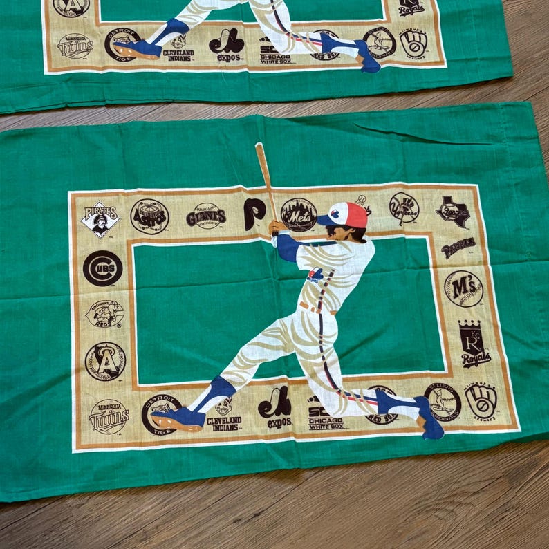 May include: Green fabric with a baseball player swinging a bat. The fabric has a border of gold and brown baseball team logos, including the Pirates, Mets, Cubs, and Royals.