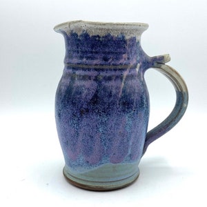 May include: A handmade ceramic jug with a light blue and purple glaze. The jug has a rounded body and a handle.