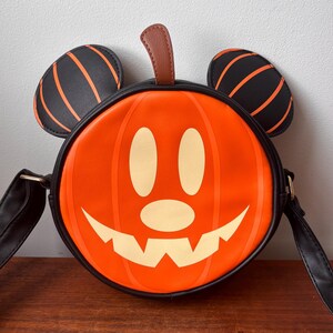May include: Orange pumpkin-shaped crossbody bag with a smiling jack-o'-lantern face. The bag features black Mickey Mouse ears with orange stripes and a brown stem. The bag has a black strap and trim, perfect for Halloween.
