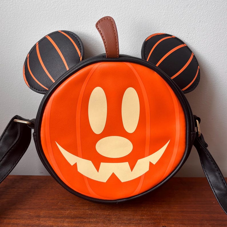 May include: Orange pumpkin-shaped crossbody bag with a smiling jack-o'-lantern face. The bag features black Mickey Mouse ears with orange stripes and a brown stem. The bag has a black strap and trim, perfect for Halloween.