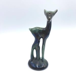 May include: A ceramic deer figurine with a glossy finish. The deer is primarily dark green and blue, with a small, round base. The figurine has a long neck and slender legs, and the ears are pointed upwards.