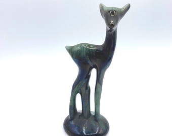 May include: A ceramic deer figurine with a glossy finish. The deer is primarily dark green and blue, with a small, round base. The figurine has a long neck and slender legs, and the ears are pointed upwards.