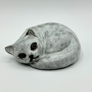 Puede incluir: Una escultura de cerámica de un gato durmiendo. El gato está enroscado en una bola con la cabeza apoyada en las patas. La escultura está pintada en color gris claro con detalles marrones para los ojos y la nariz.
