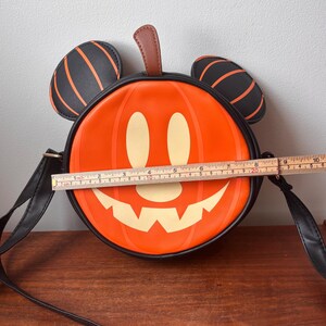 May include: Orange and black Halloween-themed crossbody bag shaped like a pumpkin with a smiling jack-o'-lantern face. The bag has black Mickey Mouse ears with orange stripes and measures approximately 8 inches in diameter.