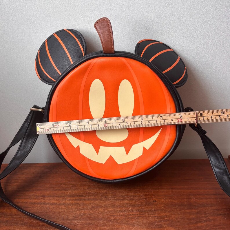 May include: Orange and black Halloween-themed crossbody bag shaped like a pumpkin with a smiling jack-o'-lantern face. The bag has black Mickey Mouse ears with orange stripes and measures approximately 8 inches in diameter.