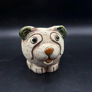 May include: A ceramic figurine of a dog with brown, white and green glaze. The dog has a friendly expression with large eyes and a wide smile.