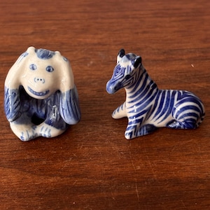 Peut inclure: Deux petites figurines en céramique : un singe bleu et blanc se couvrant les oreilles et un zèbre bleu et blanc couché. Les figurines sont sur une surface en bois. Le singe a un visage souriant et le zèbre a des rayures bleues.