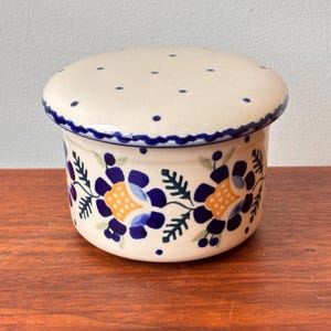 Pode incluir: Uma manteigueira de cerâmica com tampa. O prato cilíndrico tem uma base e tampa creme. Apresenta um padrão floral azul com detalhes amarelos. A borda e a tampa são decoradas com pontos azuis e uma borda azul ondulada.