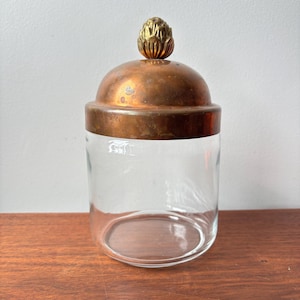 Pode incluir: Um frasco de vidro transparente com tampa cor de cobre e um adorno decorativo dourado. O frasco está sobre uma superfície de madeira. A tampa tem formato de cúpula e uma faixa de cobre ao redor da borda.
